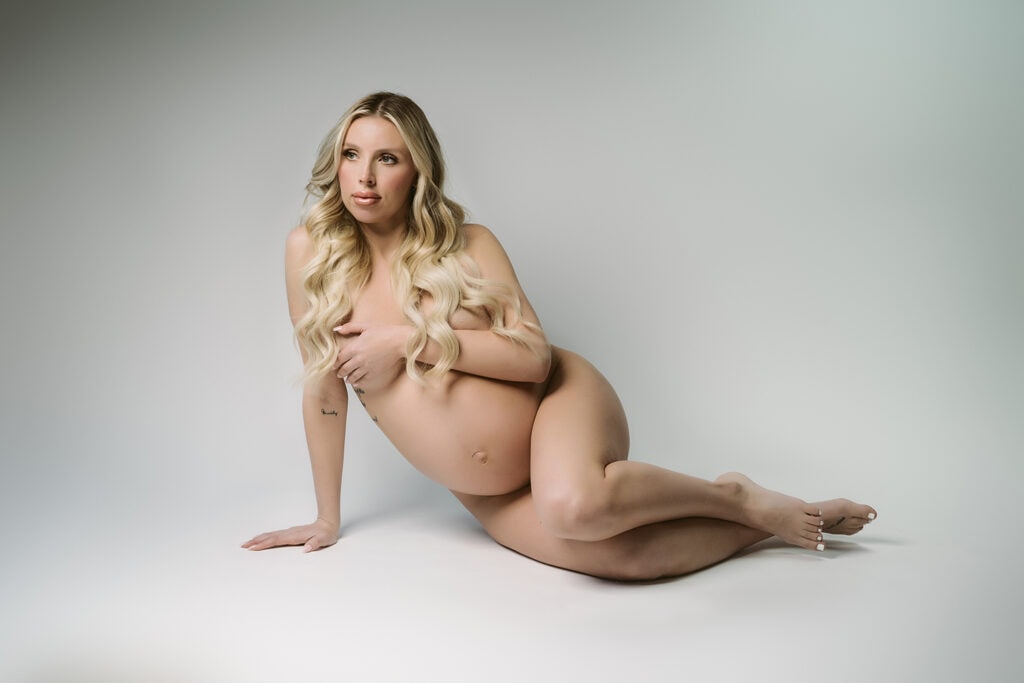 nude maternity photo, blonde woman, pregnant, laying on her side against a white backdrop propped up on one hand.